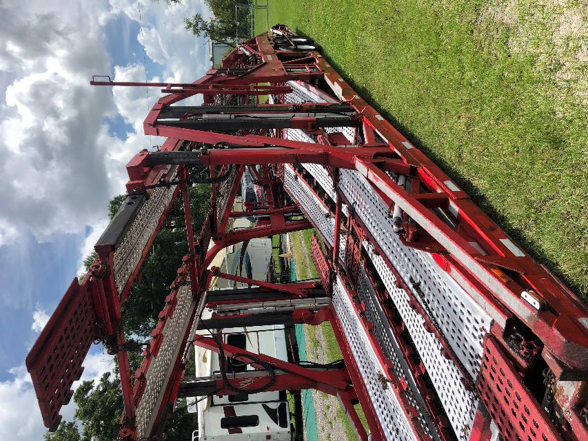 Truck carrier trailer with multiple car hauler racks and metal perforated ramps, red frame, parked on grass.