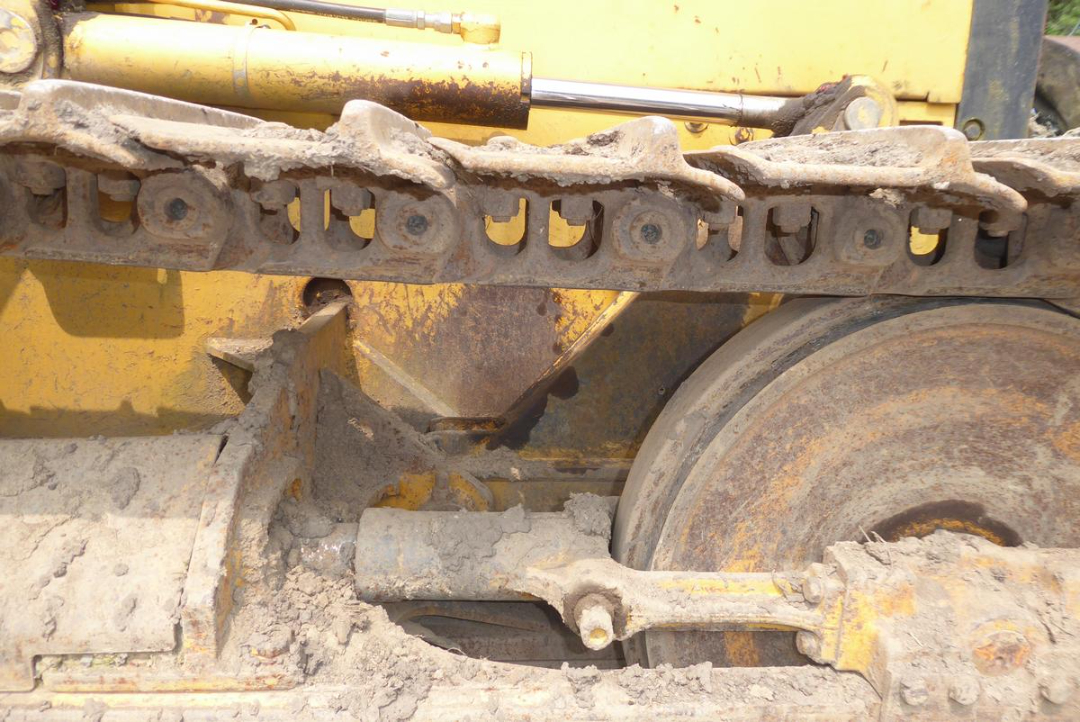 Side view of a tracked dozer undercarriage with track links, rollers, and wheel; dusty yellow machinery visible.