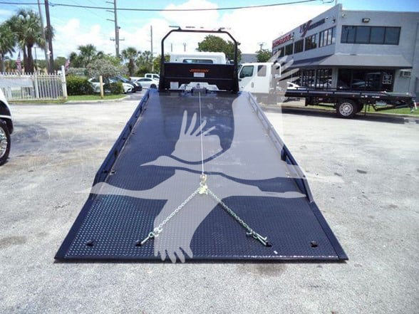 Flatbed tow truck ramp with safety chains and a winch point, seen from the end, with a building and vehicles in the background.