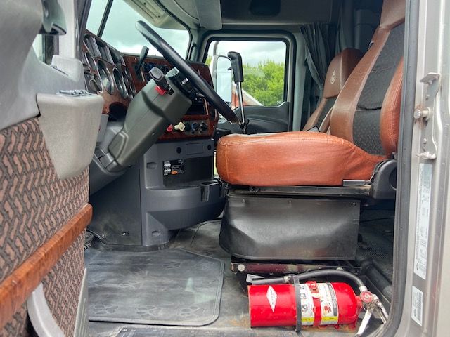Inside view of a 2010 Mack CXU613 conventional sleeper truck cab showing brown leather driver's seat, dashboard with wood trim, steering wheel, and a red fire extinguisher on the floor near the door.