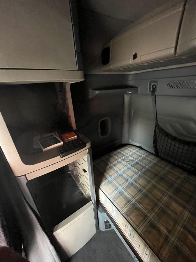 Cab interior of a 2015 Freightliner Cascadia sleeper showing a plaid mattress, overhead storage, and a small cabinet with a keypad-like device.