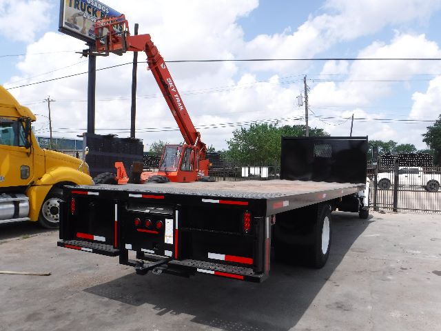 Flatbed truck with long steel deck and black rear chassis, shown from rear-right in a lot with other trucks and a yellow cab nearby.