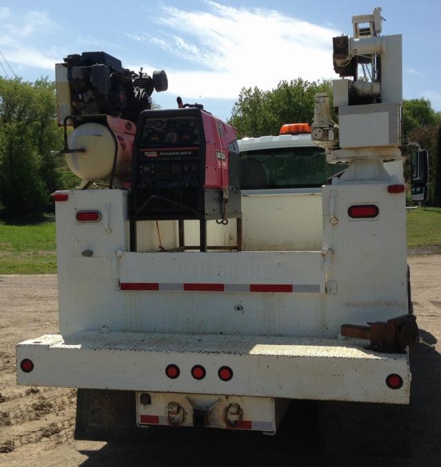 Truck bed with mounted generator and hydraulic equipment, rear view, white utility truck, engine/generator on left, boom/crane apparatus on right, tail lights and bumper visible.