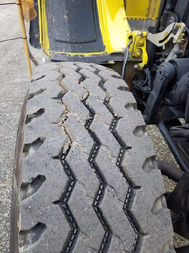 Truck tire close-up with deep tread and wheel well; yellow cab and suspension components visible in background.