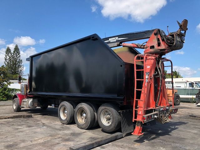 2013 Peterbilt 388 grapple truck with a large black dump body and crane on a trailer at a yard.