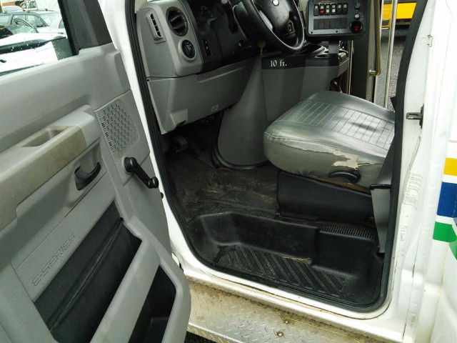 Ford E-450 front cab interior showing gray driver's seat, steering wheel, and dashboard with control panel; door sill area and ribbed floor mat visible.