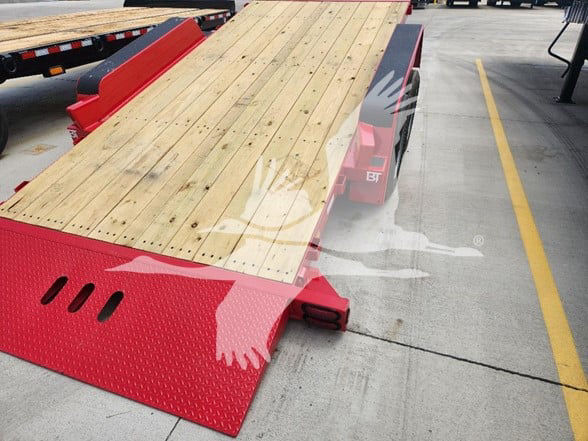 Behnke flatbed trailer with wooden deck and red gooseneck/fender area. Visible: red metal ramp with diamond plate texture, wood planks on deck, black wheel well, BT logo on fender.