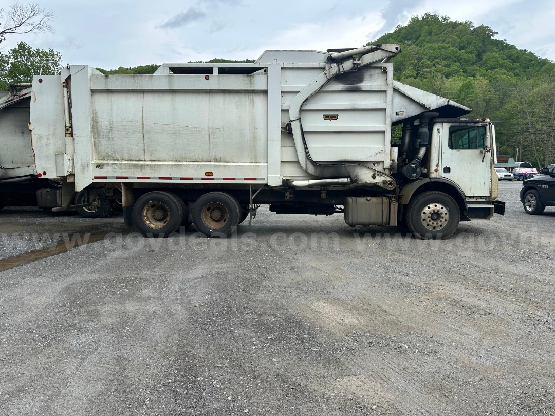 Truck side view showing a white Mack MRU613 dump truck with large dumping bed and hydraulic arm.