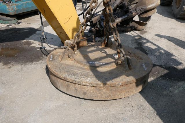 Circular magnet weight plate with lifting chains attached to a yellow excavator arm. Weathered metal surface with small white stencils or markings.