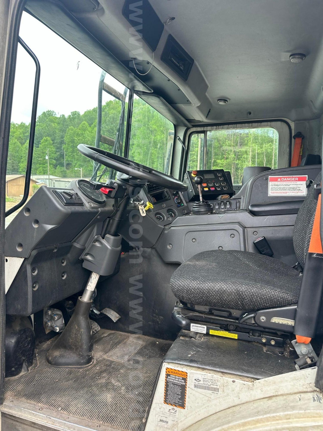 Truck cab interior with steering wheel, gear lever, control panel, and driver seat from left side of cab. Visible are dashboard controls and a warning label near the seat base.