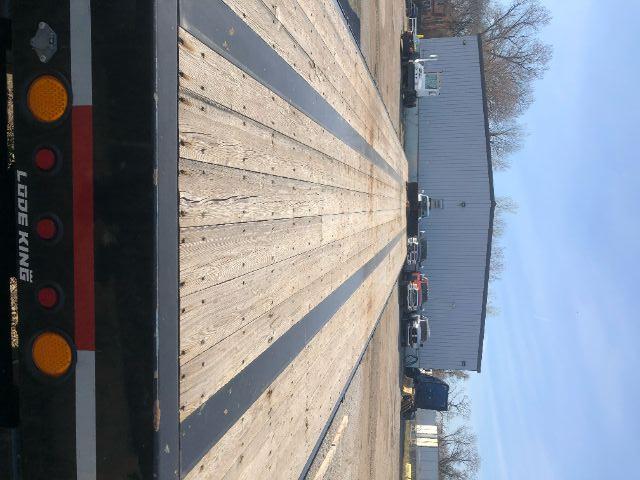 Load King PF 948 oilfield flat deck trailer: long wooden deck with black frame, rear taillights and orange reflectors, industrial building in background.