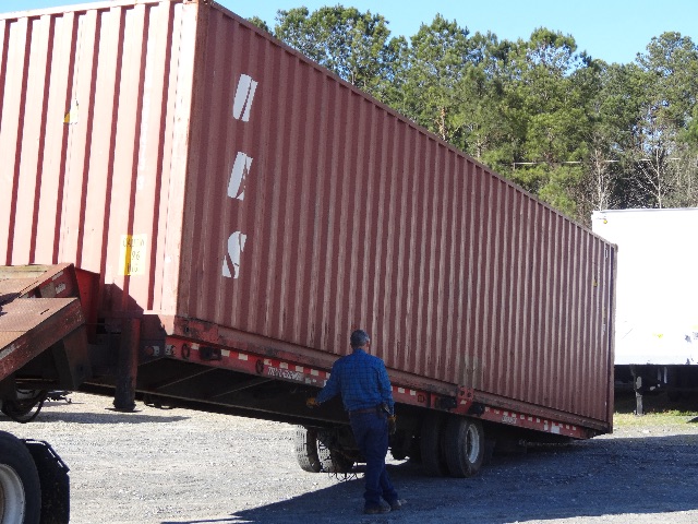 Used red cargo container mounted on trailer with a person standing beside, side view showing corrugated metal walls and white markings.