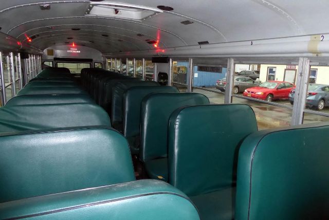 School bus interior with teal vinyl seats, double-occupancy rows, windows along both sides, overhead ceiling with lights, and exit sign at far end.