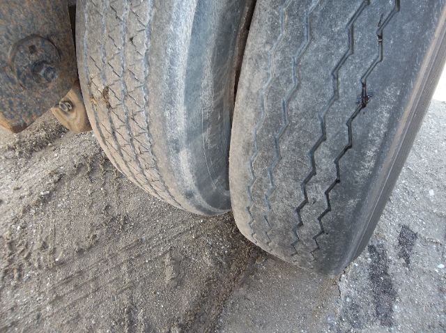 Tandem trailer axles with worn tires and dirt on the ground.