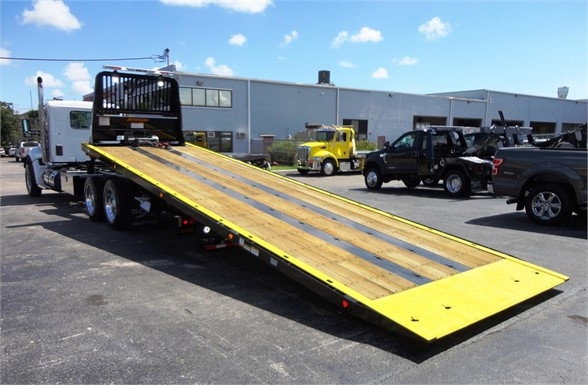 Kenworth W900 flatbed tow truck with a long yellow and wood-plank ramp extending from the rear; rear view shows wheel-laden trailer and equipment yard.