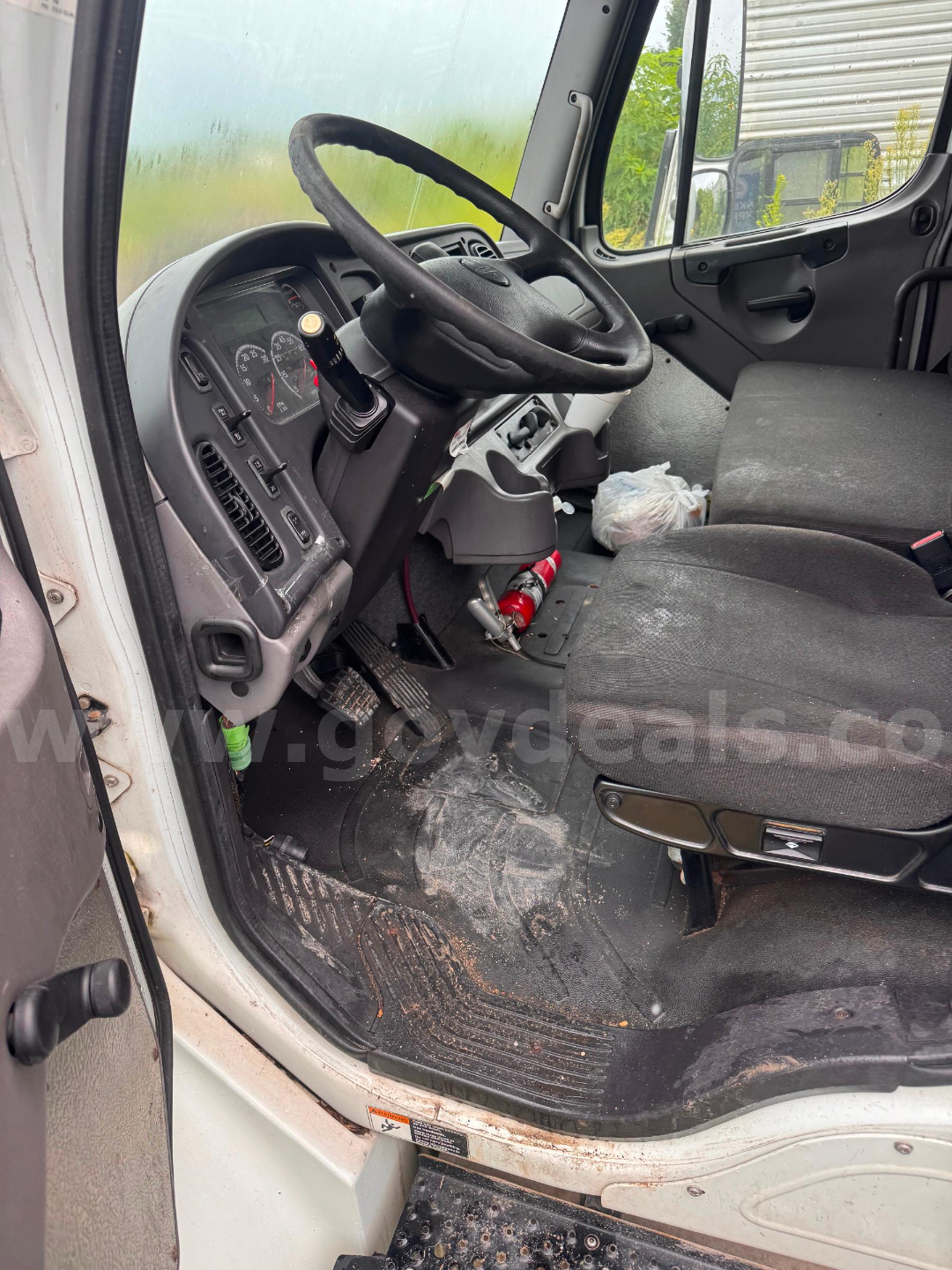 Truck cab interior showing steering wheel, dashboard gauges, and driver seat of a Freightliner M2 106, with floor mat and some items on the floor.