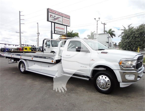 Tow truck with flatbed, white Ram 5500 HD SLT, visible decals and a dealership sign in background.