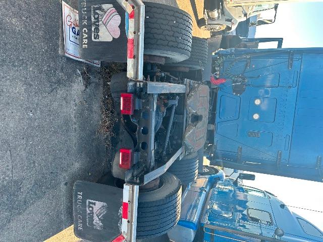Image shows the rear of a blue semi truck with dual wheels and a flatbed trailer attachment. Visible text includes 'RUL CARE' on a license or tag and a small sign attached to the trailer frame. There are taillights and metal support beams.