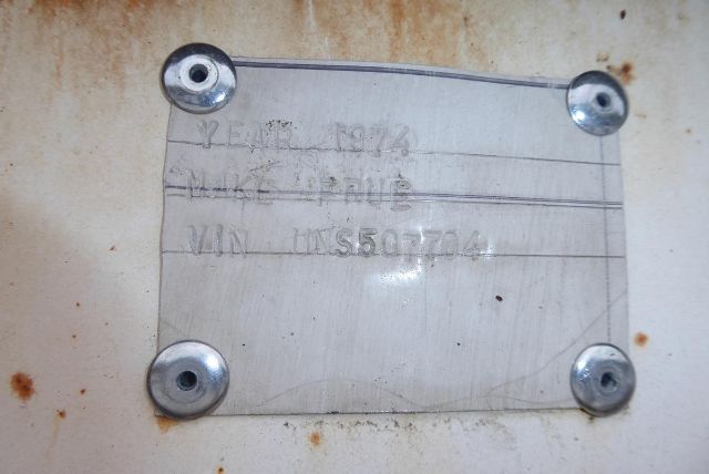 Pale metal identification plate on a trailer wall with embossed text: 'EAC 1972' and 'VIN: UNS507704' with four screws in each corner.