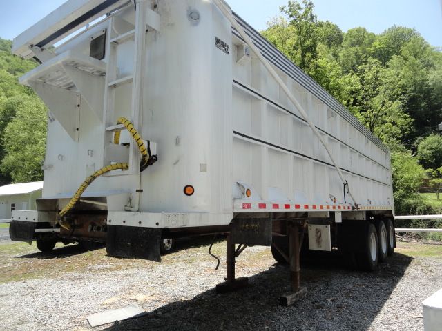 Rear-left side of a white dump trailer with hydraulic hoses and ramps; shows landing gear and underside. No visible text readable from equipment labels.