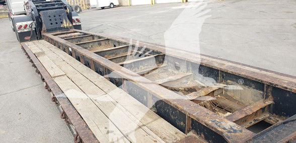Trailer frame of a Fontaine 55 Ton lowboy with wooden deck planks and steel I-beam rails; visible rust and wear along the deck; some equipment in background.