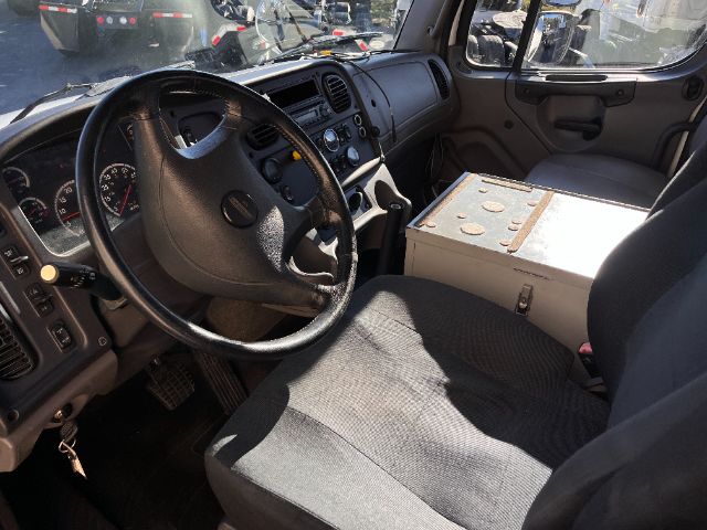 Cab interior of a 2015 Freightliner M2 truck showing steering wheel, dashboard, and a metal toolbox/passenger seat area.
