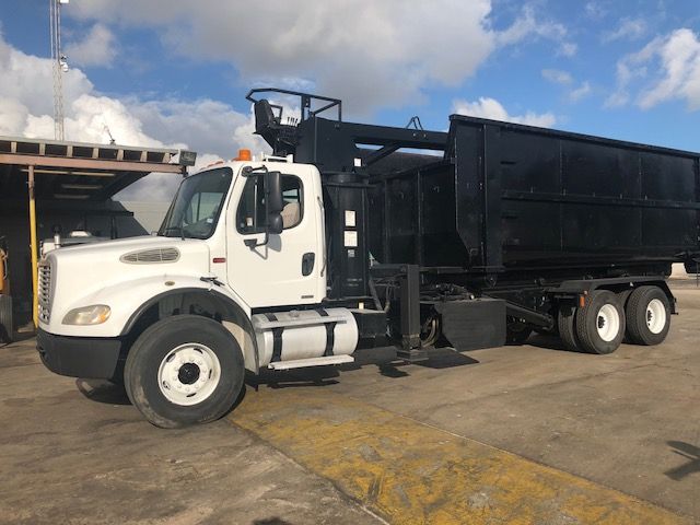 White Freightliner M2-112 grapple truck with black dump body, side view highlighting cab and front wheel, used in heavy machinery handling.