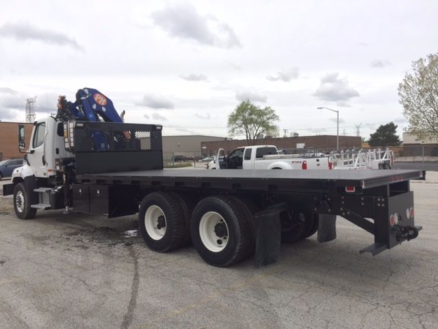 Flatbed truck with knuckle boom crane mounted on cab and long black flatbed, three axles, white wheels, rear extended flat surface and equipment rack near cab.