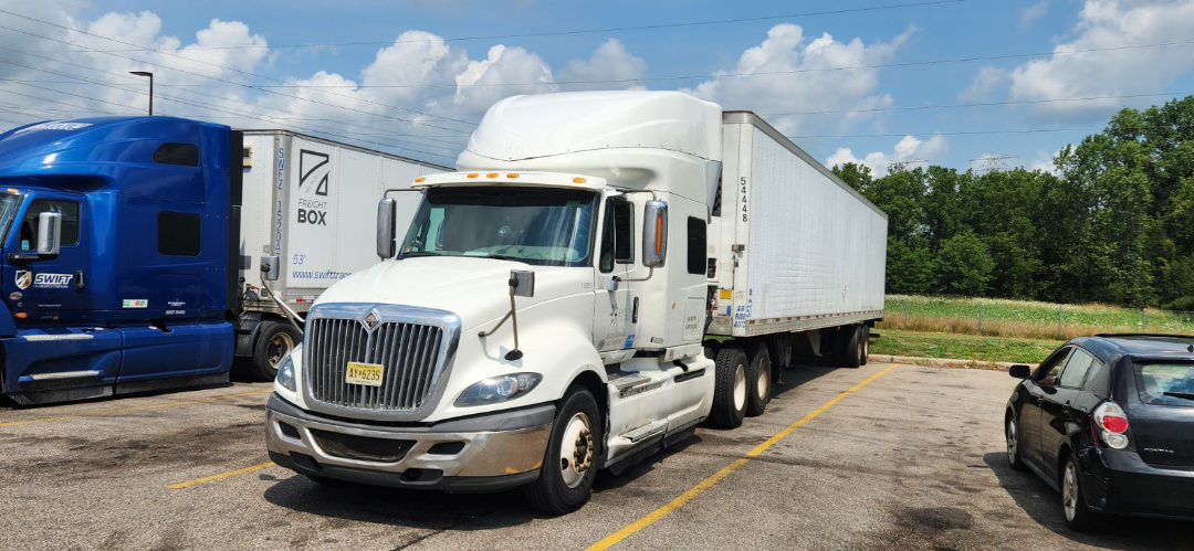 White 2016 International ProStar tractor with a long white trailer, parked in a lot with a blue truck on the left and a black car on the right. Clear sky with some clouds.