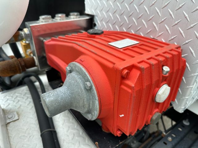 Red pump/drive unit mounted on a diamond plate surface with bolts and a gray input shaft, part of a GMC W5500 truck system.