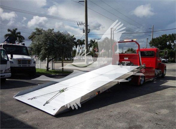 Peterbilt 337 tow truck with a long, elevated flatbed in the bed area, red cab, white ramp, and other equipment visible in a parking lot.
