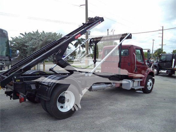 Freightliner Business Class M2 106 refuse truck chassis showing a hoist/loader arm on the rear with maroon cab and white wheels.
