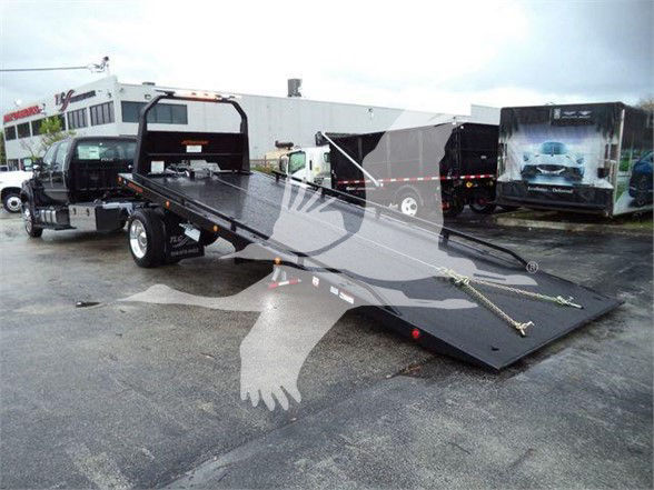 Ford F-750 Tow Truck with a lowered flatbed ramp and dual-wheel rear. Visible text includes dealership branding on the side doors and background trucks; VIN and stock numbers not legible on image itself.