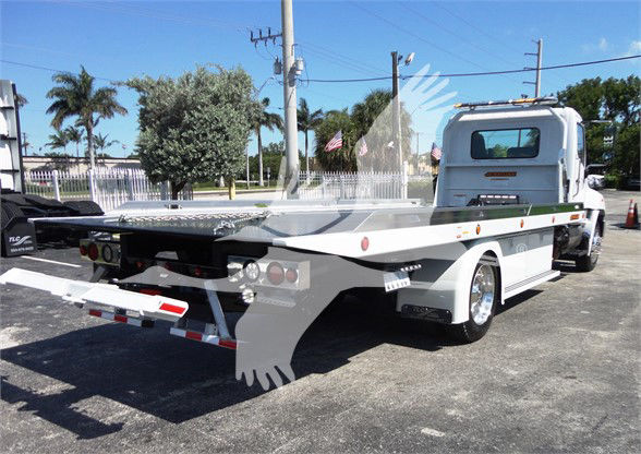 Flatbed tow truck with a white cab and long flatbed, rear view showing loading ramps and taillights. Stock area and blue sky.