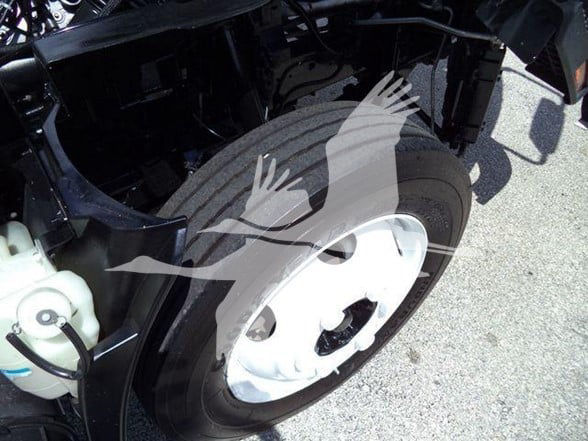 Isuzu NPR HD truck front wheel well with white wheel and black tire showing part of chassis; reservoir bottle visible in left foreground.