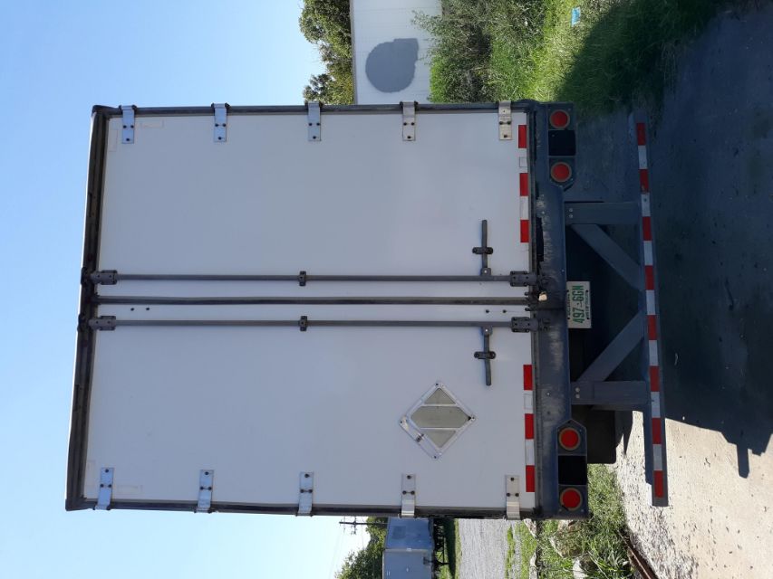 Rear view of a white utility trailer with double doors, hinges, and reflective red and white tape along the lower edge.