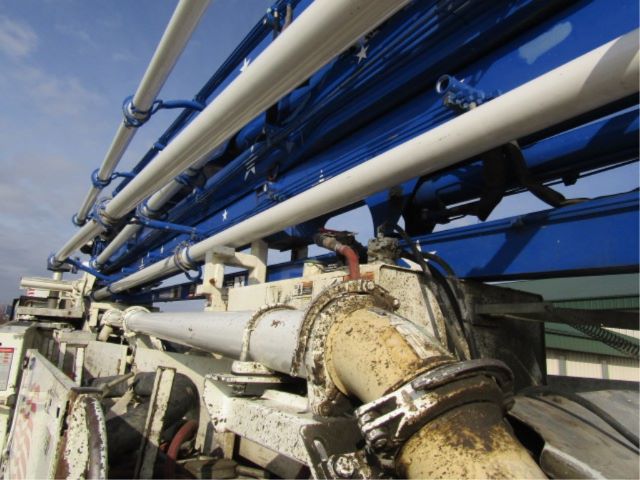 Farmed blue Mack T-series truck component with hydraulic hoses and mechanical joints; close-up of white and blue piping and rusty hydraulic gear housing.