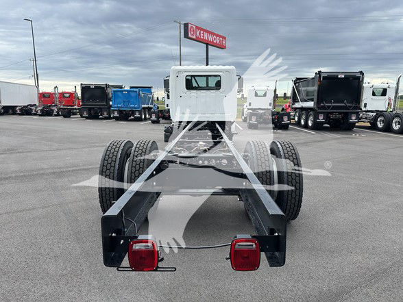 Rear view of a cab and chassis truck frame with dual rear wheels, taillights, and a small white cab in the distance; Kenworth dealership lot in background.