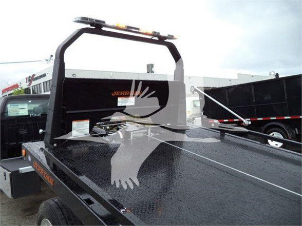 Flatbed tow truck bed with a menacing arching frame and a rear black metal surface, small orange warning sticker near the front, and a light bar on top; no legible branding visible.
