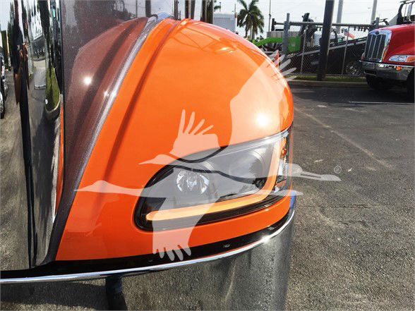 Peterbilt truck front end close-up showing orange hood and headlight, reflection on bumper, and gravel lot in background.