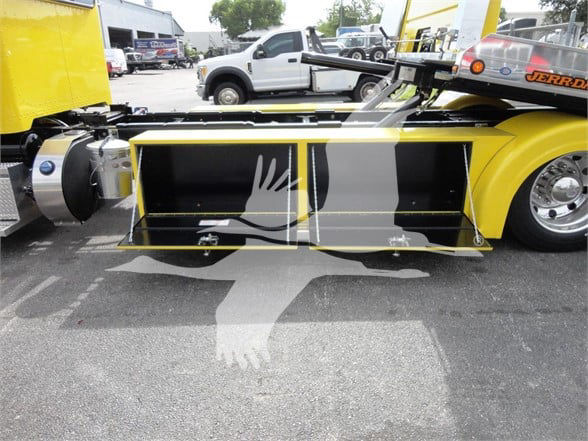 Truck side view showing yellow Peterbilt 337 tow truck with hydraulic flatbed and storage compartments; visible black storage boxes under the bed.