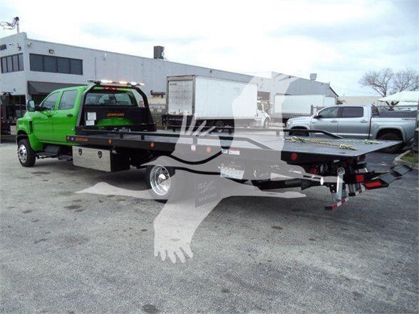 Flatbed tow truck with a bright green cab and long black flatbed, loading rear of a vehicle. There are trucks and a building in the background.