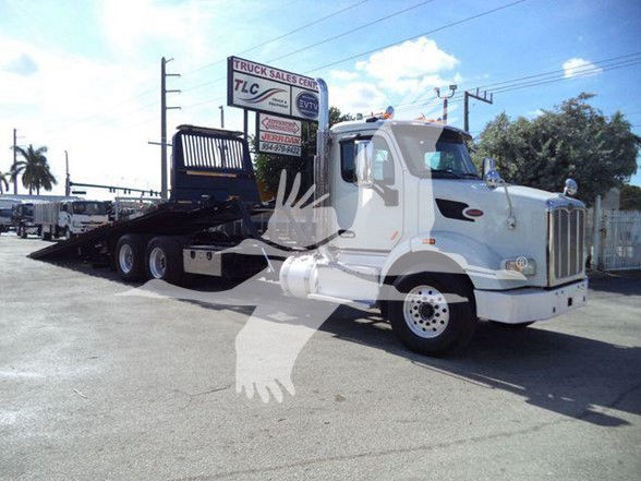 Tow truck with a white Peterbilt 567 cab and flatbed trailer, front view, on a lot with signage in background.