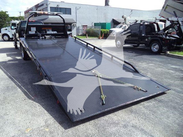 Flatbed tow truck bed with black diamond plate surface and ramps folded at rear; straps and tie-down points visible. Background shows other trucks and a building.