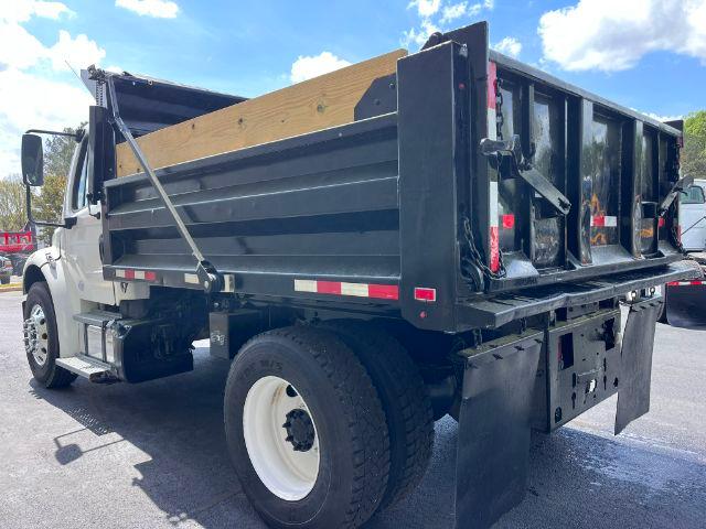 Freightliner M2 106 dump truck with black dump bed, white wheels, and wooden side railing. Side view showing tailgate area and hydraulic components.