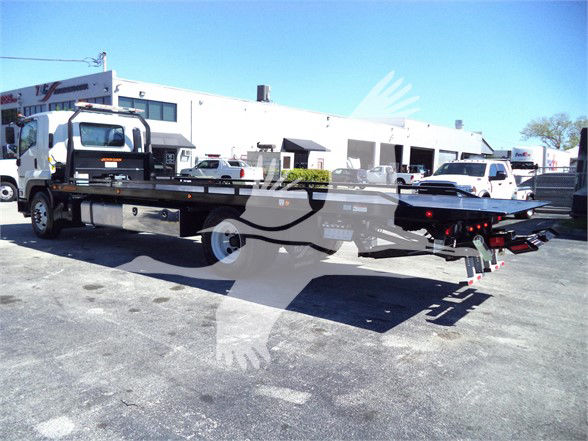 Isuzu tow truck or flatbed in a lot with dealership buildings in background. Side view shows long flatbed with wheel, ramps, and rear equipment. Visible text: none clearly legible on the vehicle itself.