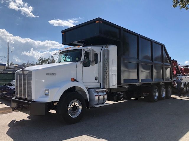 Kenworth T800 dump truck in white cab with large black dump body, side exhaust, and multiple rear axles; daytime outdoor lot.