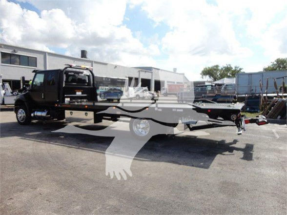 Tow truck with a black cab and a long flatbed trailer, International MV model, parked in an industrial lot with buildings in the background.