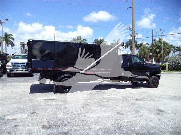 Truck with long black cargo box on a heavy-duty black cab chassis, side view, in a lot with trees and sky in background.