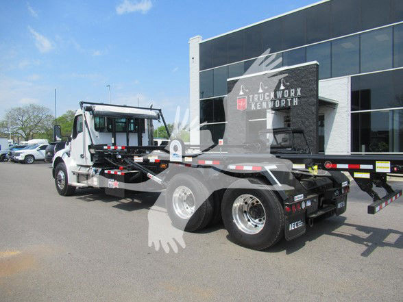 Kenworth T480 heavy-duty flatbed tow truck in a dealership lot, white cab, gray flatbed, chrome wheels, side mirrors, and dealership building in background.